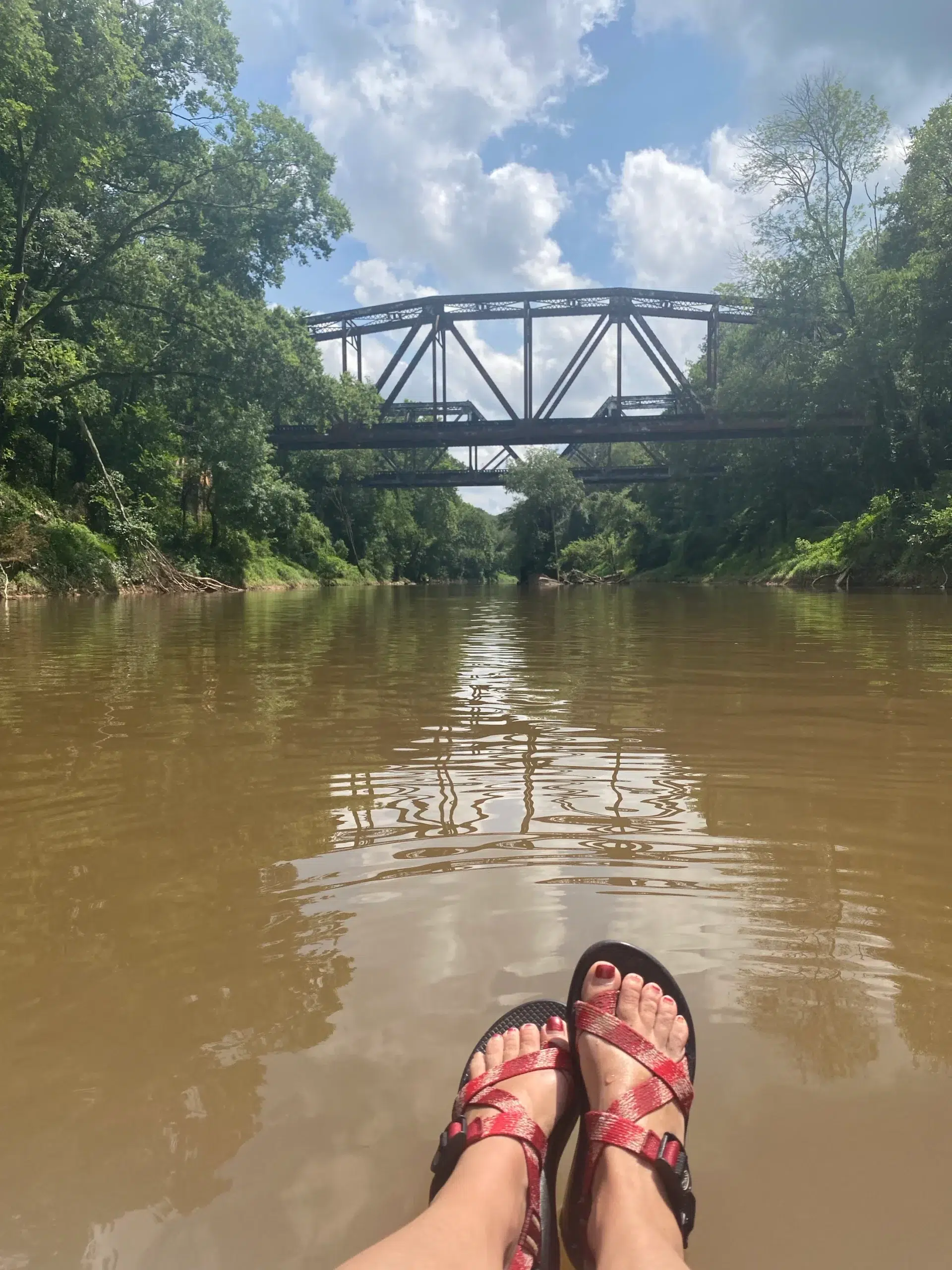 A person with sandals relaxes on a river, their feet visible in the foreground. Green trees line the riverbanks and a metal train bridge spans the water under a partly cloudy sky.