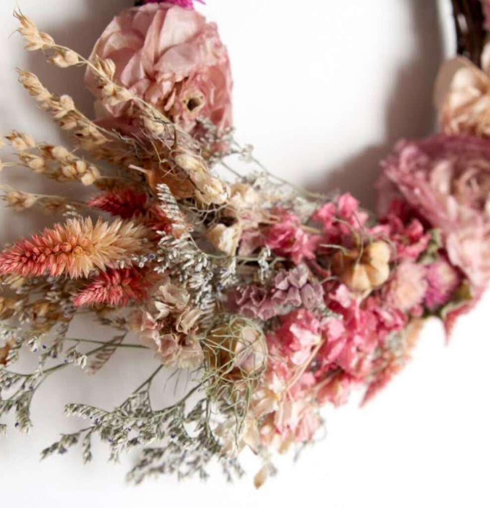 Close-up of a decorative wreath made of dried flowers, including pink roses, lavender sprigs, and wheat stalks, arranged on a white background.