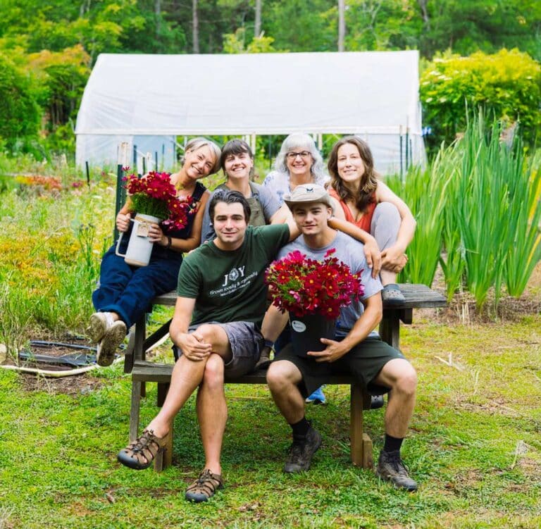 Six smiling people pose outdoors in front of a greenhouse, holding red flower bouquets. Three sit on a bench while three stand behind them. Lush greenery surrounds the group on a bright day.