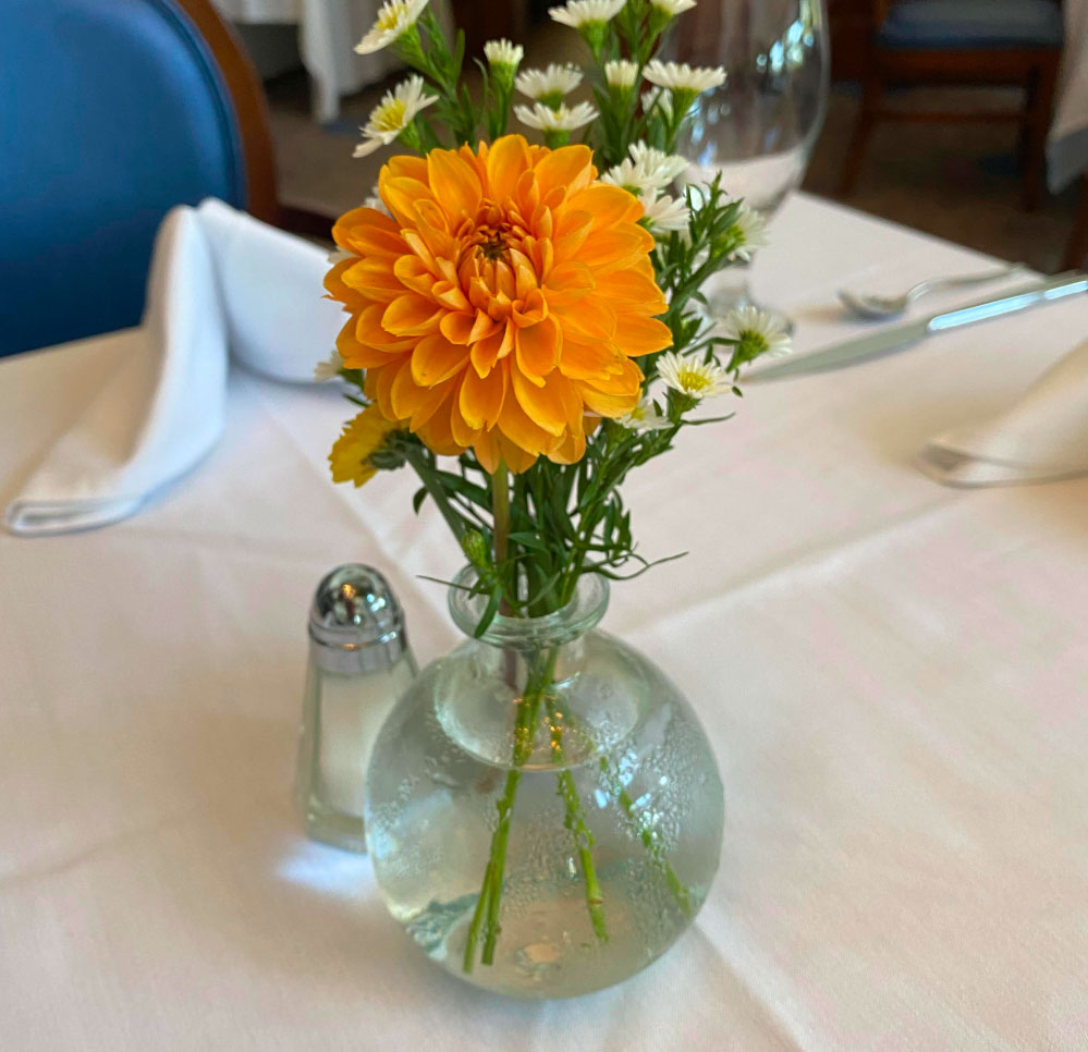 A small glass vase with an orange dahlia and white daisies sits on a white tablecloth, next to a salt shaker and neatly folded napkins in a restaurant setting.