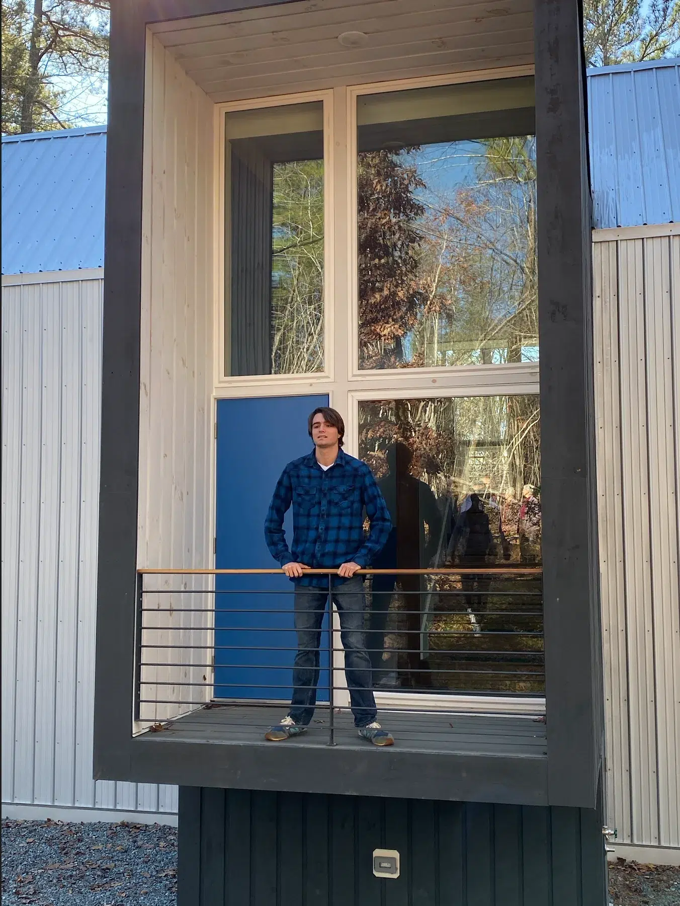 A person wearing a blue plaid shirt and jeans stands on a small balcony with a metal railing, in front of a modern building with large windows and light-colored siding. Trees are reflected in the window.