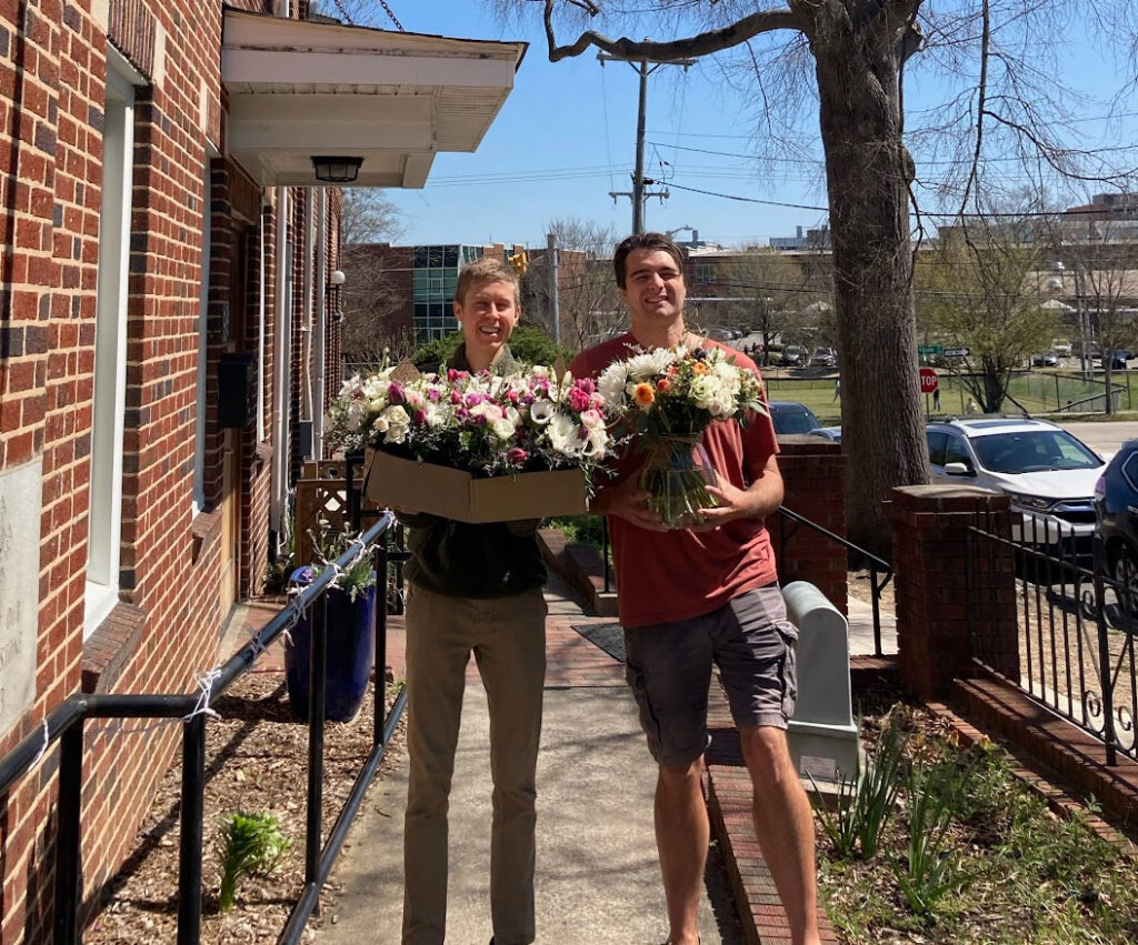 Two smiling people stand outside on a sunny day holding arrangements of colorful flowers, one with a large box and the other with a bouquet, beside a brick building and a tree.