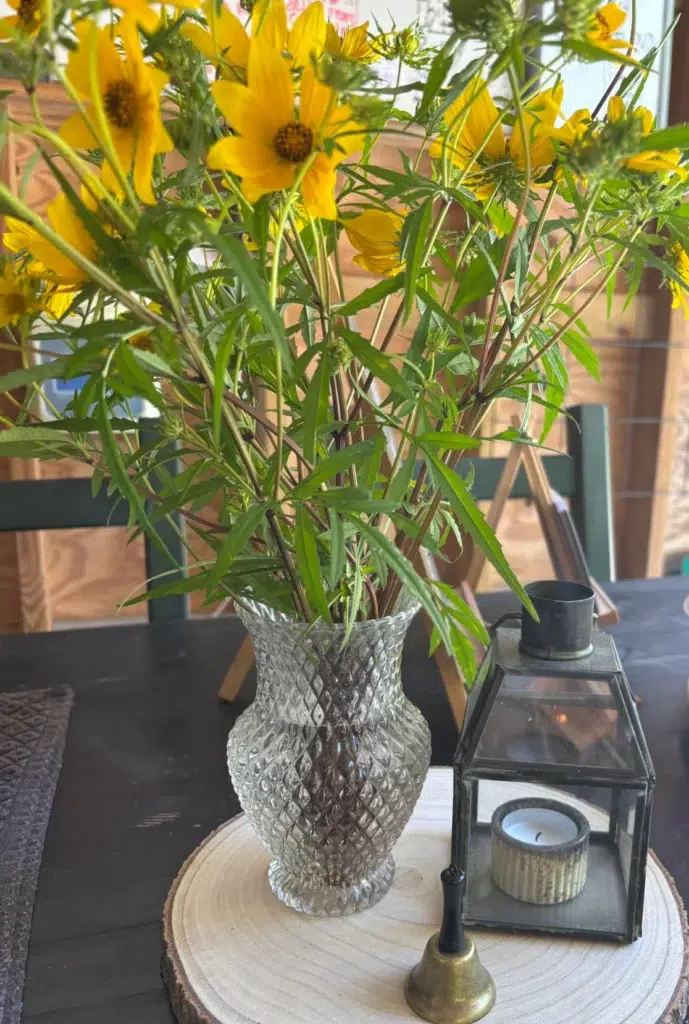 A clear, textured glass vase holds yellow flowers with green leaves on a wooden slice. Next to it is a small metal lantern with a candle and a brass candle snuffer on a dark table.