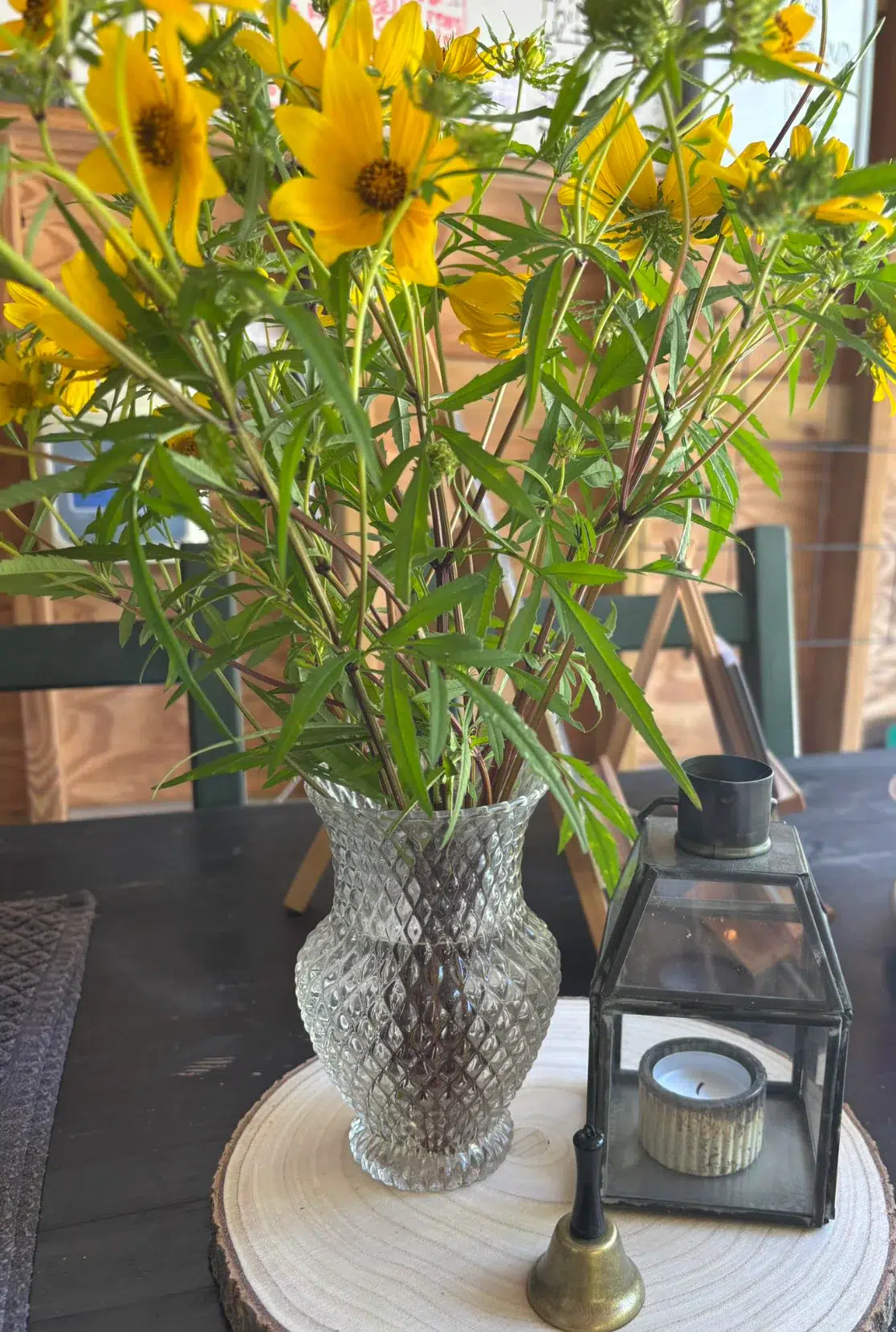 A clear, textured glass vase holds yellow flowers with green leaves on a wooden slice. Next to it is a small metal lantern with a candle and a brass candle snuffer on a dark table.