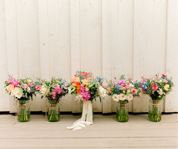 Five glass jars filled with colorful flower bouquets sit on a wooden floor in front of a white paneled wall. The middle bouquet has a white ribbon tied around the jar.