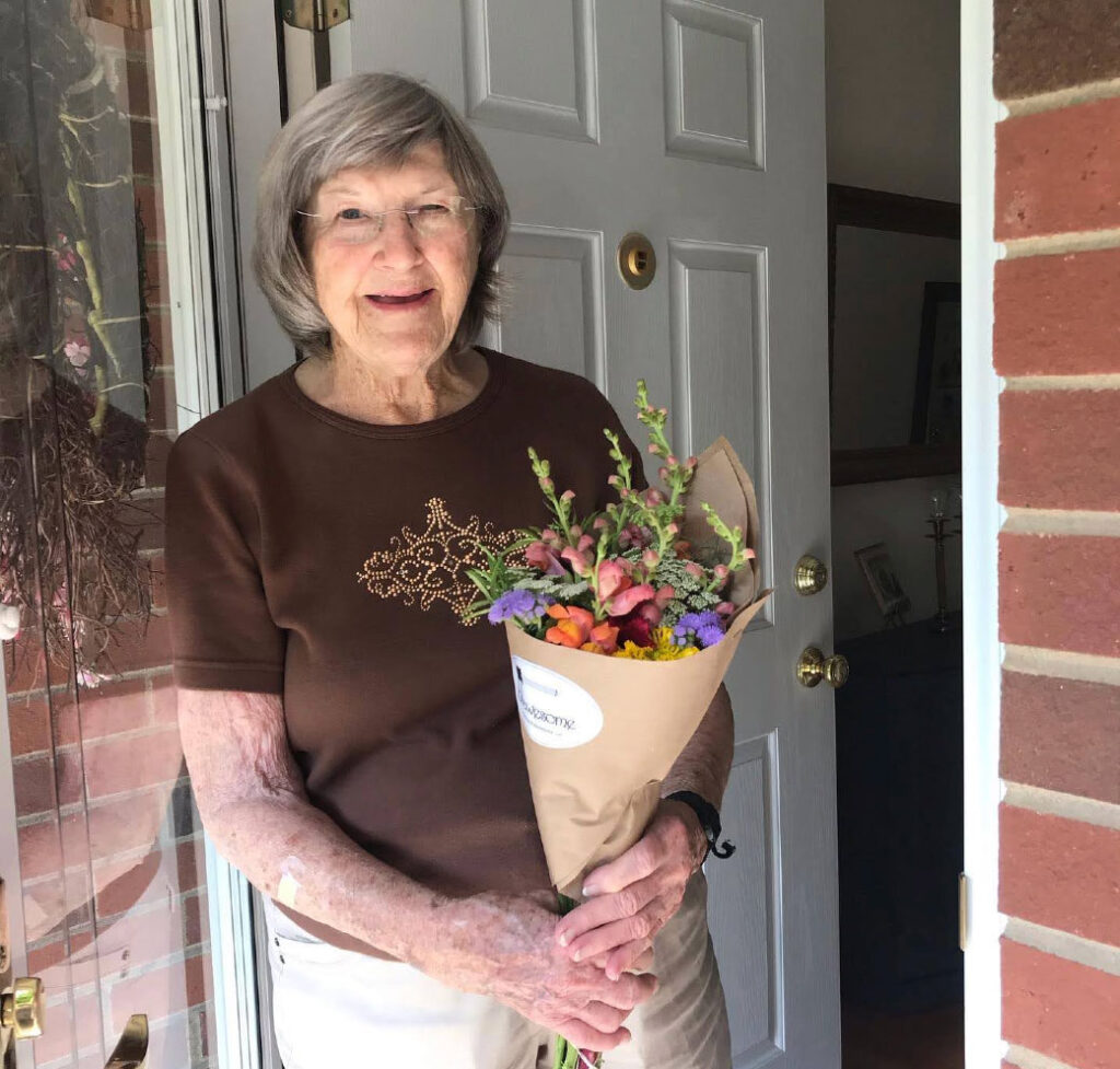 An elderly woman with gray hair and glasses stands smiling at an open door, holding a colorful bouquet of flowers wrapped in brown paper. She wears a brown shirt and light pants. Brick and part of a hallway are visible.