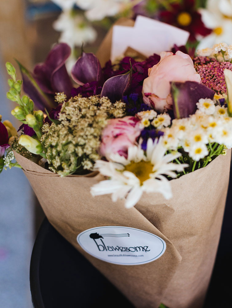 A bouquet of mixed flowers, including daisies, roses, and purple blooms, wrapped in brown paper with a sticker labeled blawesome. The bouquet is colorful and fresh.