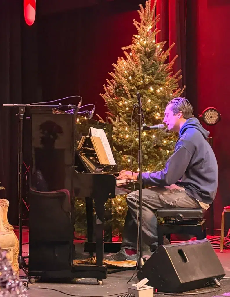 A person in a hoodie plays piano and sings into a microphone on stage, with a decorated Christmas tree lit up in the background and red curtains behind them.