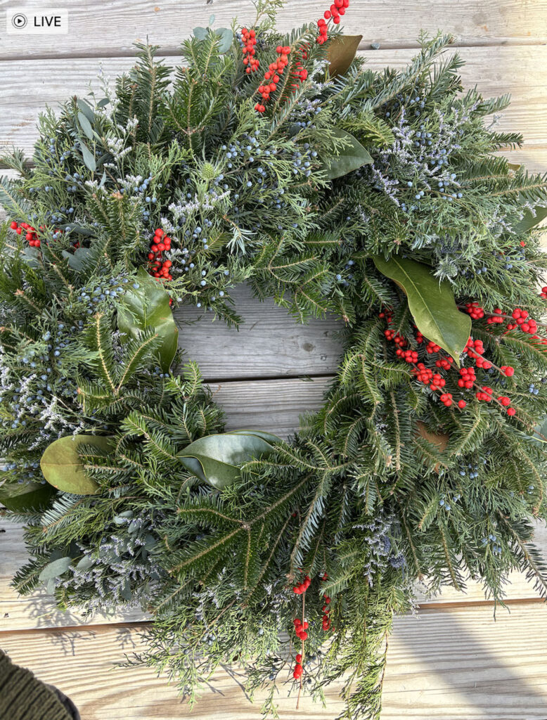 A festive wreath made of evergreen branches, red berries, and large green leaves sits on a wooden surface.