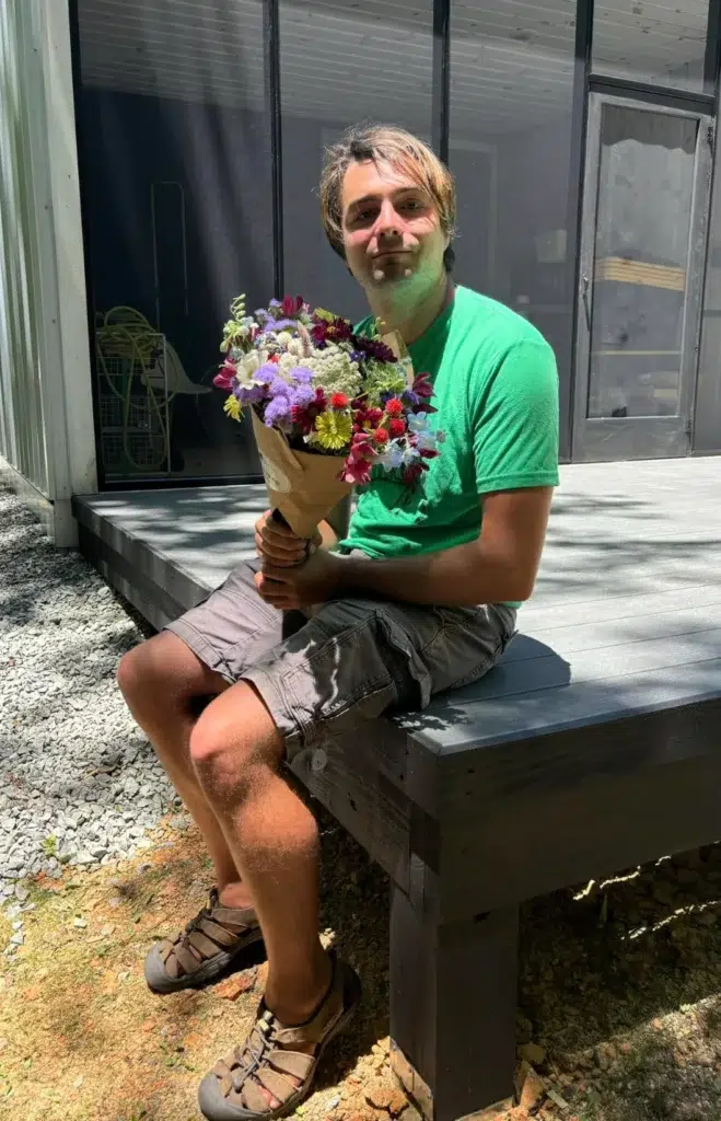 A man in a green shirt and gray shorts sits on a wooden deck holding a colorful bouquet of flowers, smiling in the sunlight. The background shows part of a house and some outdoor equipment.