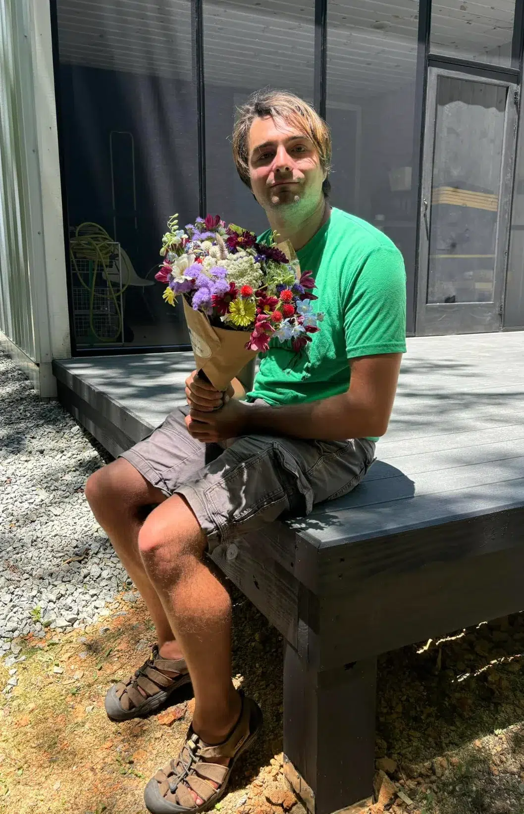 A man in a green shirt and gray shorts sits on a wooden deck holding a colorful bouquet of flowers, smiling in the sunlight. The background shows part of a house and some outdoor equipment.