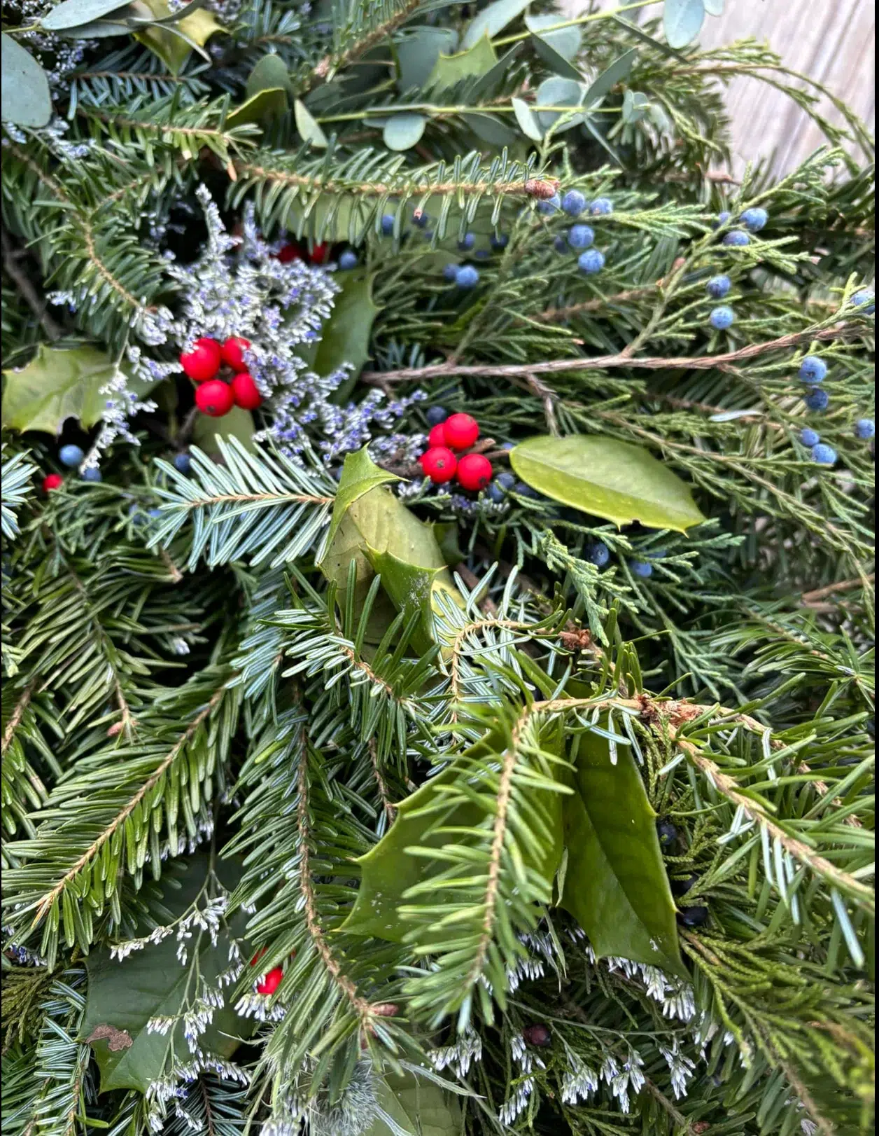 A close-up of evergreen branches with green needles, red berries, dark green holly leaves, and small clusters of blue berries, creating a festive and natural arrangement.