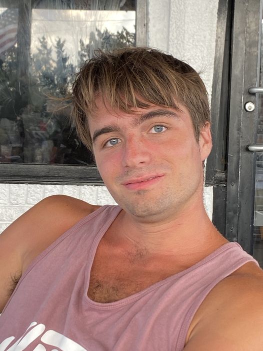 A young man with light brown hair and blue eyes is sitting and smiling. He is wearing a sleeveless pink shirt and appears relaxed. The background shows a window and a white wall.