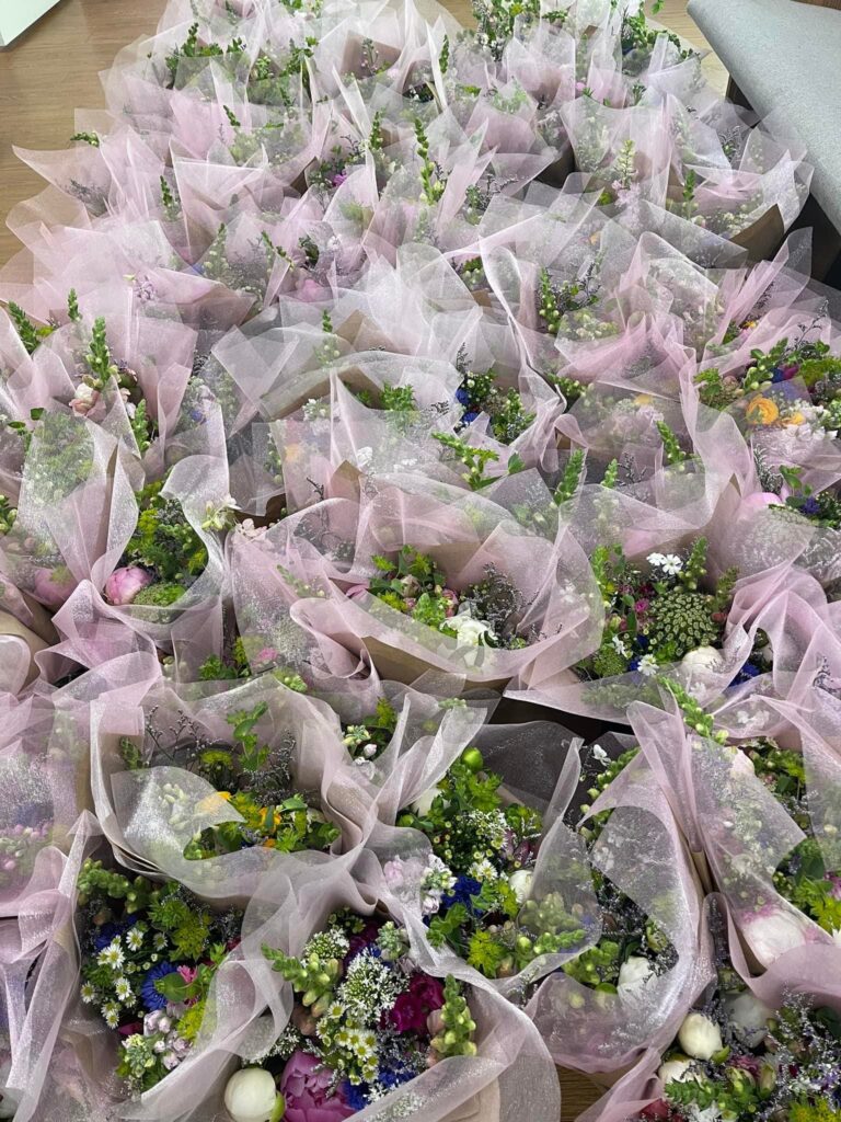 Numerous flower bouquets wrapped in light pink translucent paper are arranged closely together, featuring a mix of colorful blooms and greenery.