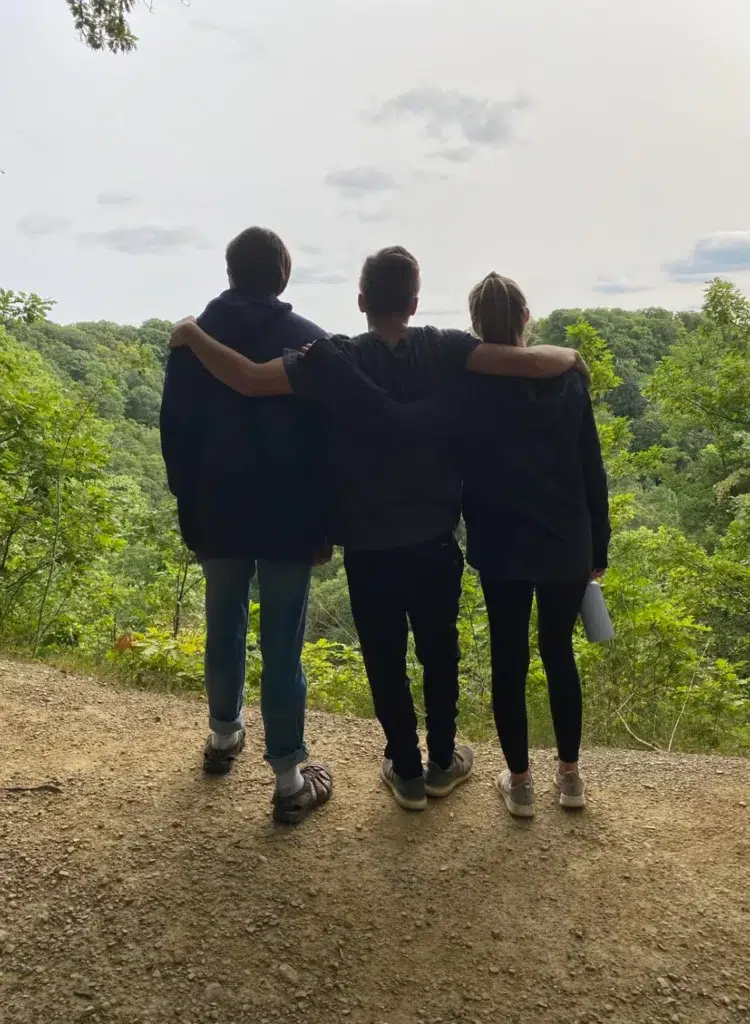 Three people stand side by side on a dirt path overlooking a green forest. The person in the middle has their arms around the other two, and all three are facing away, looking at the scenic view.