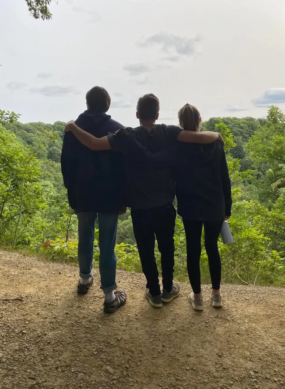 Three people stand side by side on a dirt path overlooking a green forest. The person in the middle has their arms around the other two, and all three are facing away, looking at the scenic view.