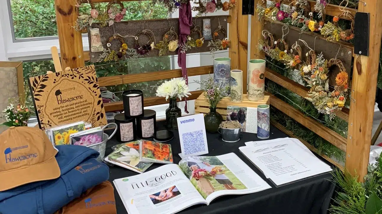 A display table featuring dried flower arrangements, candles, books, a Venmo QR code, a tan baseball cap, and brochures, with more dried flowers hanging behind on wooden racks.