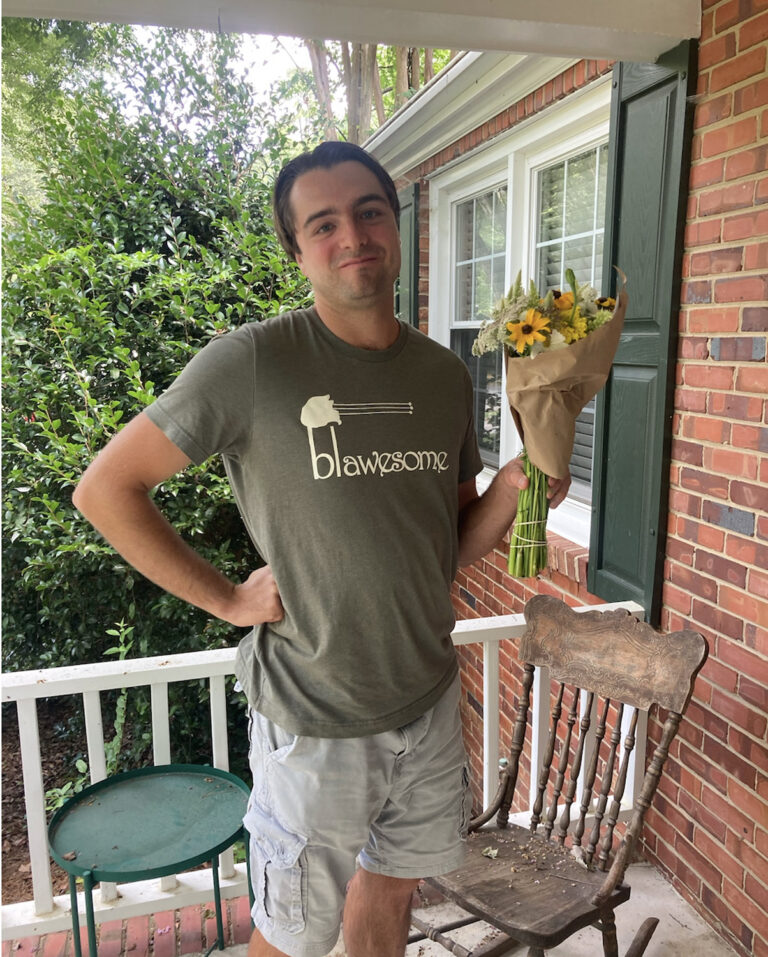 A man stands on a porch holding a bouquet of yellow and white flowers. He wears a green T-shirt that says blawsome and light shorts, and stands next to a weathered wooden chair and green table, with brick walls and greenery in the background.