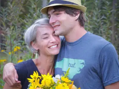 A smiling woman holding a bouquet of yellow flowers is embraced by a young man in a hat, both standing outdoors among tall greenery and yellow flowers. They look happy and close.