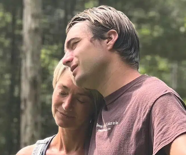 Two people stand close together outdoors, eyes closed, heads touching and leaning on each other in a moment of peaceful affection. Sunlight filters through trees in the background.