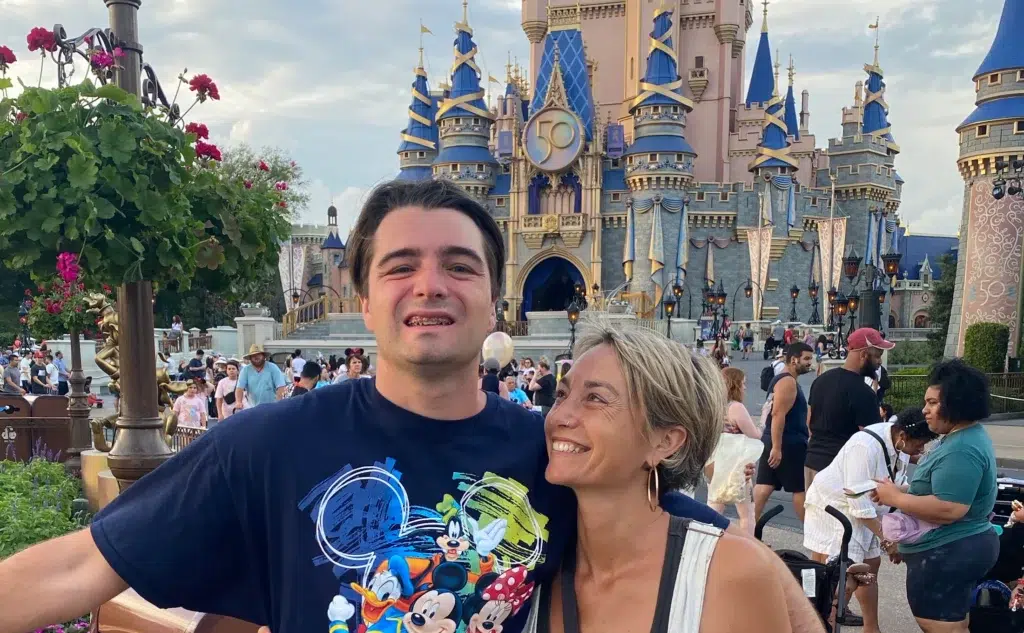 A smiling young man in a Mickey Mouse shirt stands with a woman who is looking at him fondly in front of a Disney castle, surrounded by other park visitors and colorful decorations.