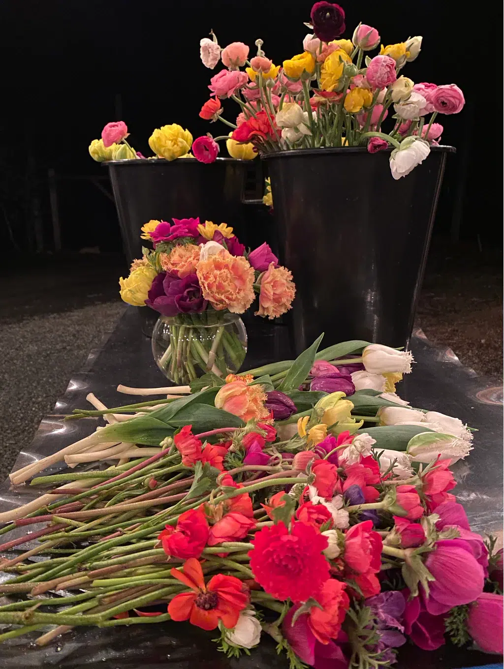 Colorful bouquets of assorted flowers, including tulips and ranunculus, are arranged in black buckets and a glass vase on a table, with more loose flowers and stems spread out in the foreground.