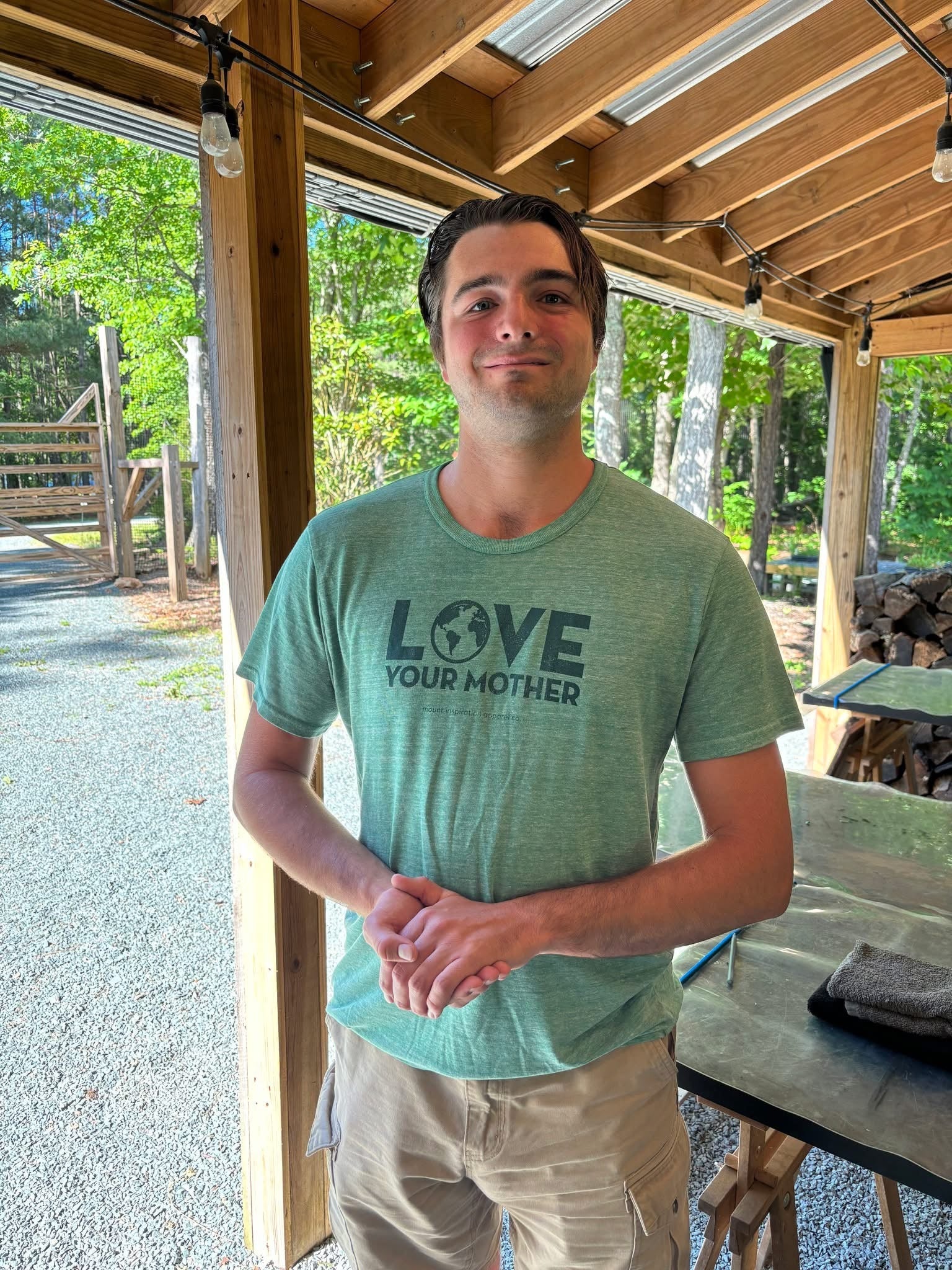 A man stands under a wooden patio, smiling at the camera. He wears a green t-shirt that says “LOVE YOUR MOTHER” with an Earth graphic and beige shorts. Sunlight filters through the trees and wooden beams around him.