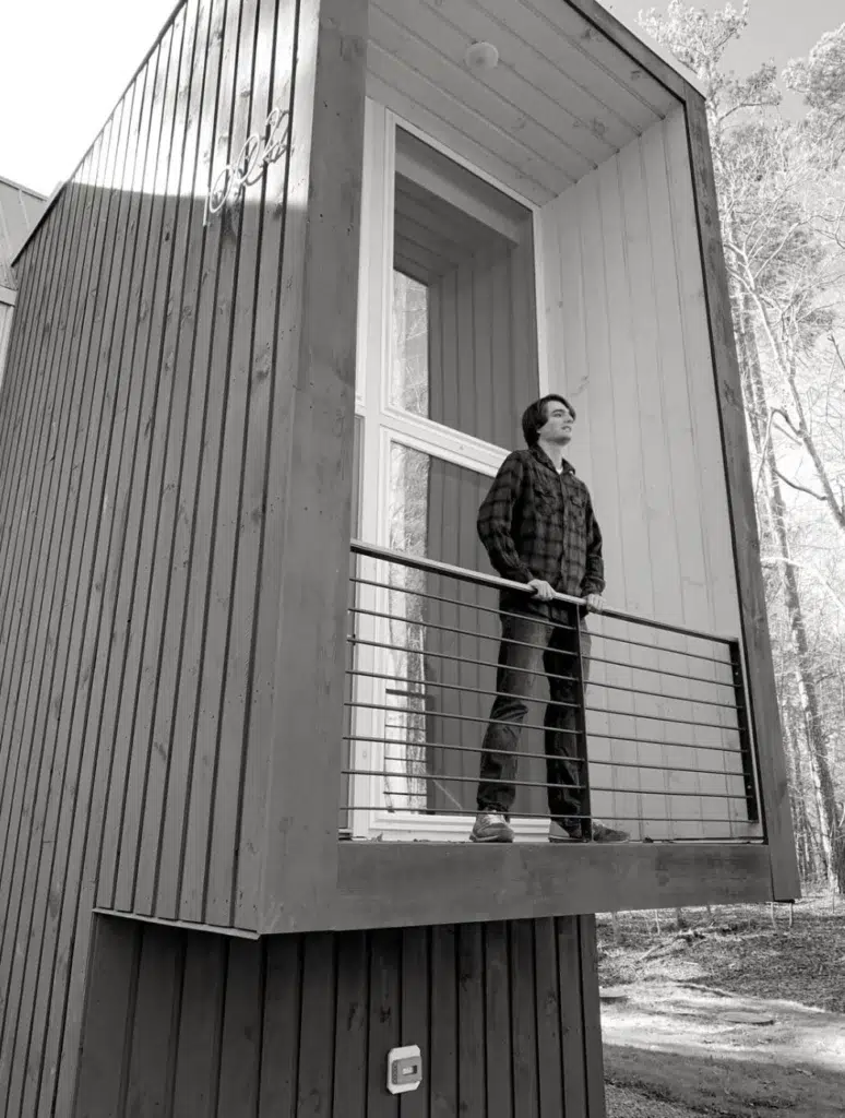 A person stands on a small, modern wooden balcony with metal railings, looking out thoughtfully. The building features clean lines and large windows, surrounded by tall trees. The image is in black and white.