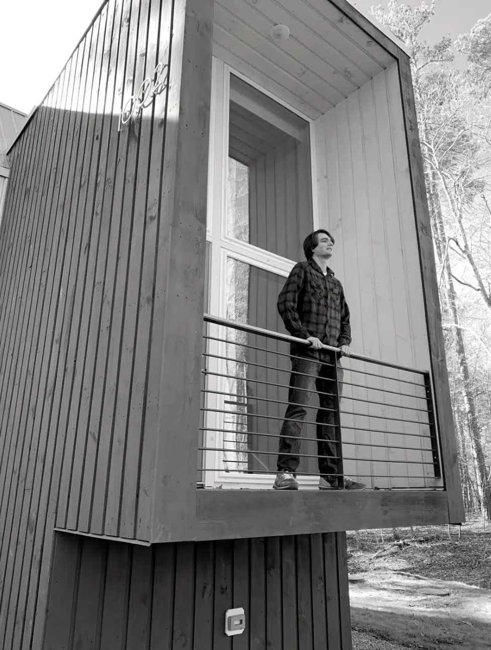 A person stands on a small, modern wooden balcony with metal railings, looking out thoughtfully. The building features clean lines and large windows, surrounded by tall trees. The image is in black and white.