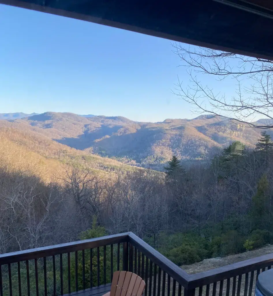 View from a deck with a wooden railing, overlooking rolling mountains covered in trees, some with bare branches, under a clear blue sky on a sunny day.