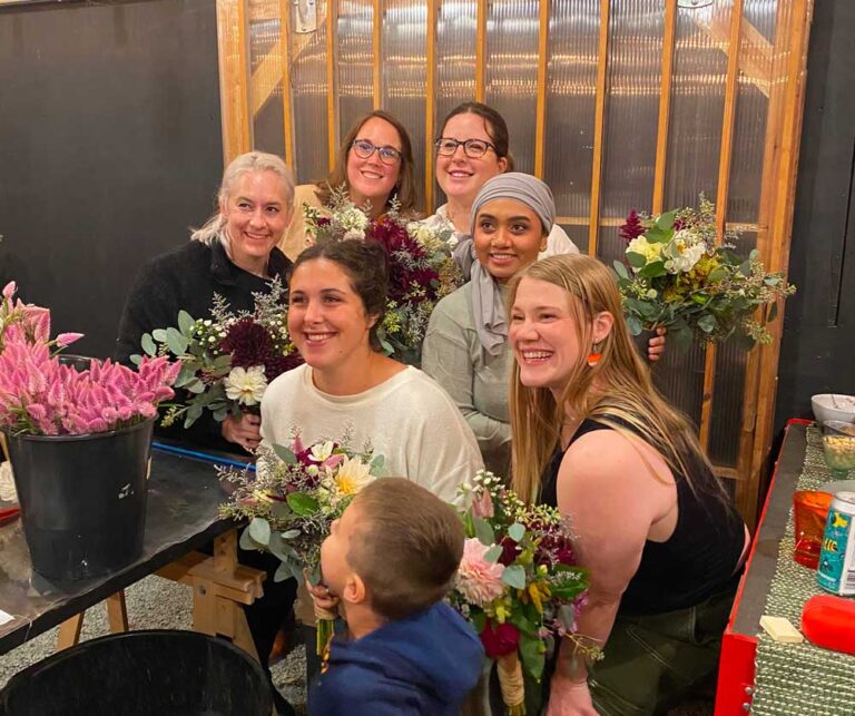 Six smiling women pose together indoors, each holding a bouquet of flowers, with a young child in the foreground. The group is surrounded by floral arrangements and appears to be enjoying a creative activity.