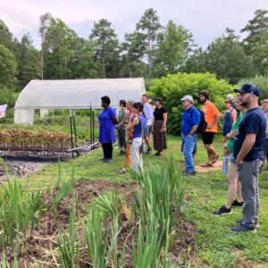 A group of people listens to a guide speaking in a community garden with raised beds, rows of plants, and a small greenhouse, surrounded by trees and greenery on a sunny day.