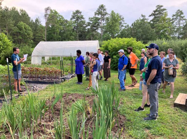 A group of people listens to a guide speaking in a community garden with raised beds, rows of plants, and a small greenhouse, surrounded by trees and greenery on a sunny day.