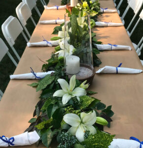 A long table with white chairs is decorated with a lush green and white floral centerpiece, featuring lilies, greenery, candles, and neatly rolled napkins tied with blue ribbon.