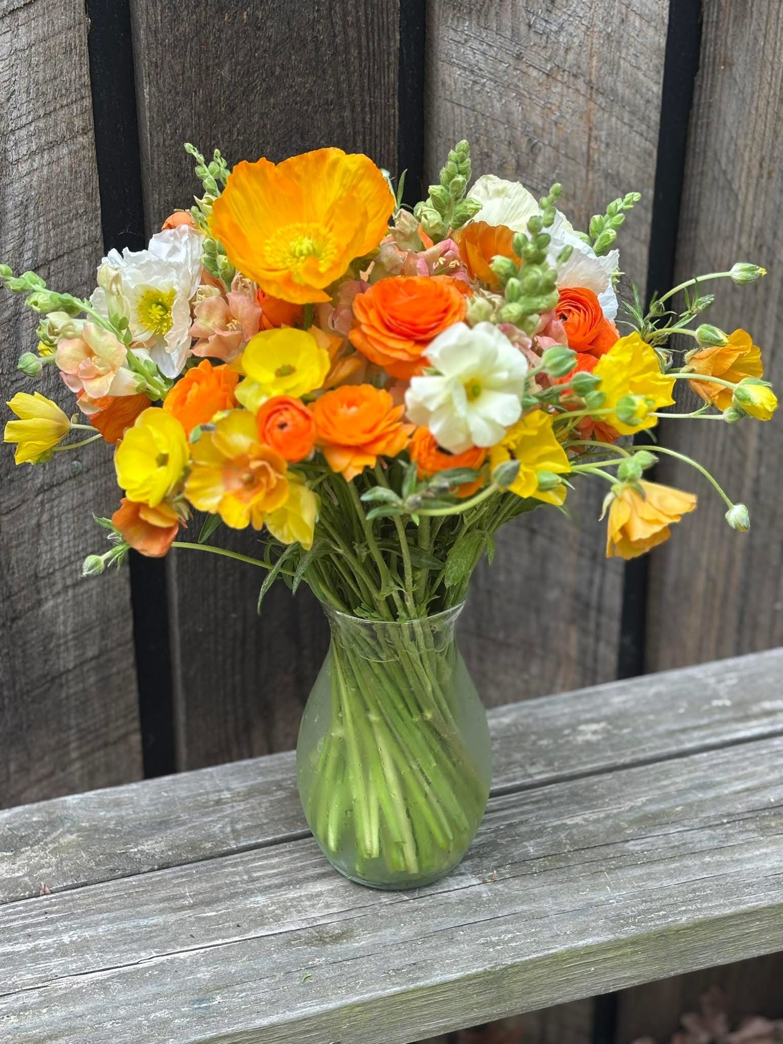 A glass vase holds a bouquet of orange, yellow, and white flowers with green stems, placed on a weathered wooden bench in front of a rustic wooden fence.