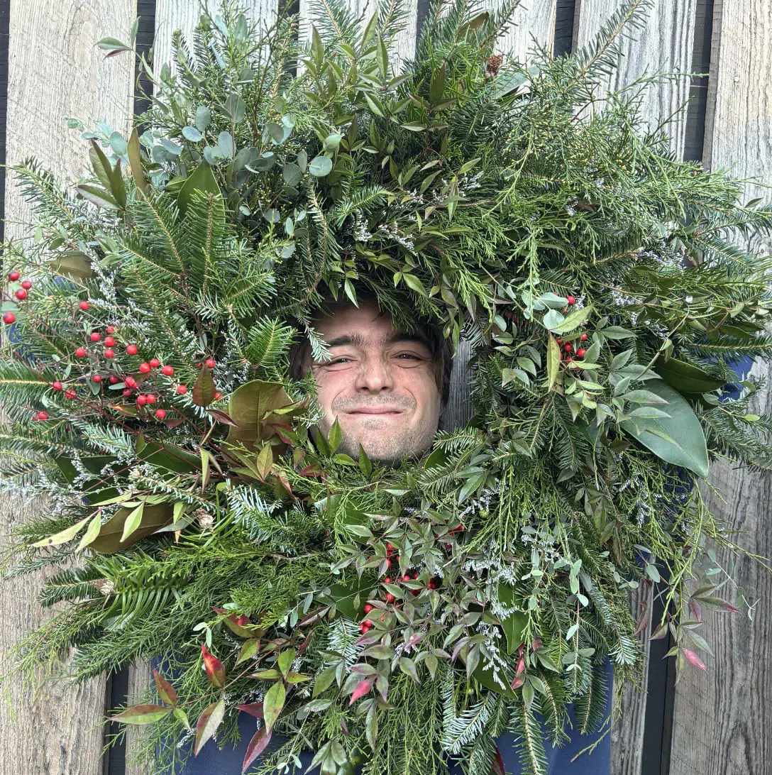 A man smiles with his face poking through the center of a large, festive wreath made of green foliage, red berries, and various leaves, set against a wooden background.