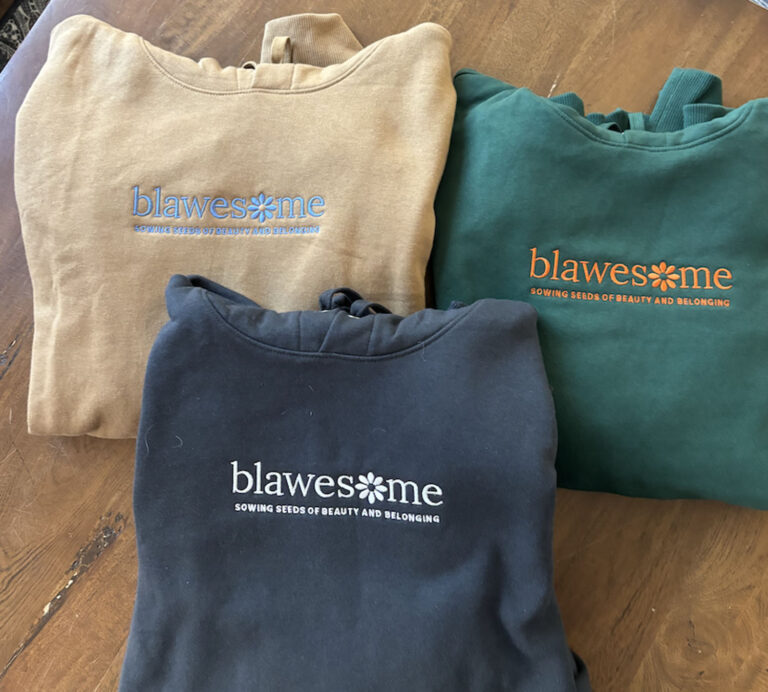 Three Blawesome Hoodies in tan, green, and black are folded on a wooden surface. Each displays "blawesome" and the slogan "sowing seeds of beauty and belonging" in colorful text.