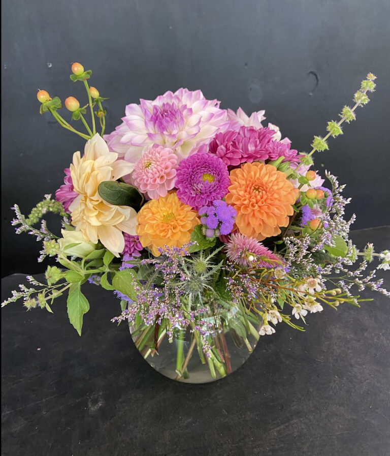 A Bubble Bowl holds a vibrant bouquet of dahlias, asters, and greenery in pink, orange, purple, and cream hues, displayed against a dark background.
