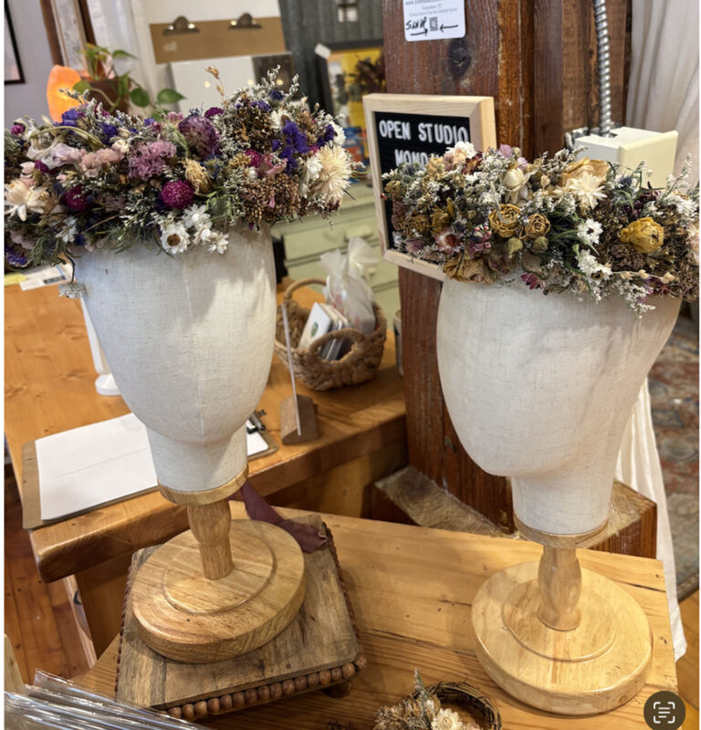Two white mannequin heads on wooden stands display the Dried Flower Crown in a cozy shop with wooden counters and assorted craft items in the background.