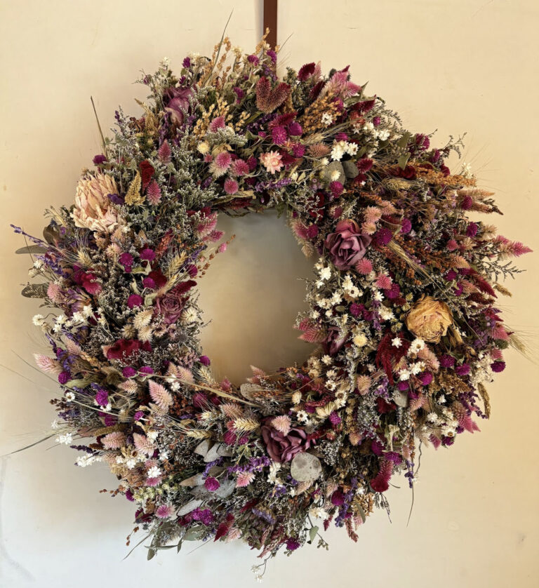 The 12"-14" Dried Floral Wreath features purple, pink, cream, and green dried flowers in a circular arrangement, displayed on a plain beige wall.