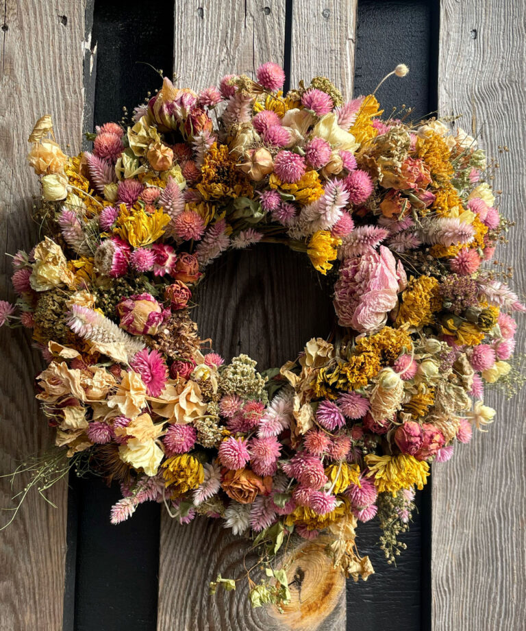 The 8" Dried Floral Wreath, featuring pink, yellow, and beige dried flowers, is displayed on a rustic wooden background with vertical planks in various tones.