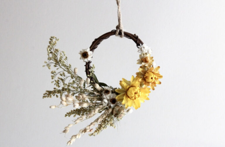 The 3" Dried Floral Wreath is a small, circular twig wreath adorned with dried yellow flowers, white blossoms, and green foliage, suspended by twine against a plain white background.