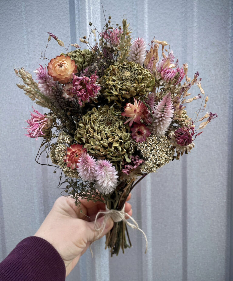A hand holds the Dried Floral Bouquet, showcasing pink, peach, and green dried flowers tied with twine. The background is a gray corrugated metal surface.