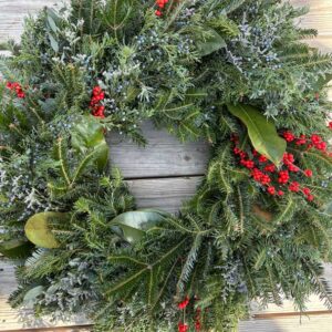 The Evergreen Wreaths-12" features pine branches, green leaves, and red berries, beautifully arranged and displayed on a wooden surface.