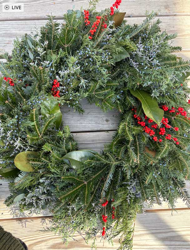 The Evergreen Wreaths-12" features pine branches, green leaves, and red berries, beautifully arranged and displayed on a wooden surface.