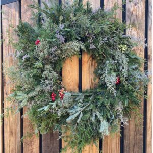 An 18" Evergreen Wreath, crafted from assorted pine branches and adorned with red berry clusters, hangs on a wooden plank wall.