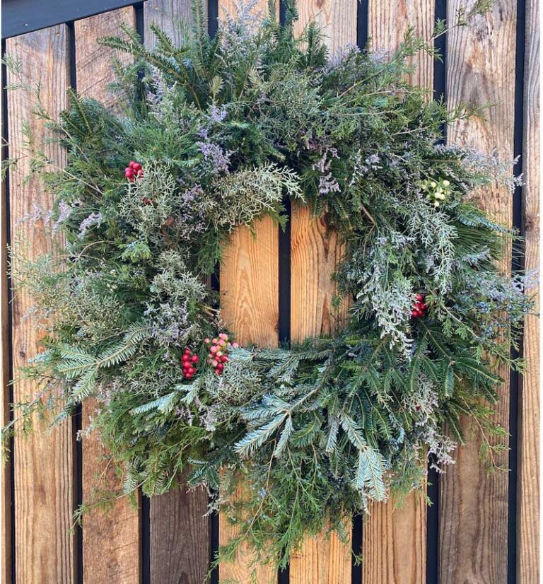 An 18" Evergreen Wreath, crafted from assorted pine branches and adorned with red berry clusters, hangs on a wooden plank wall.