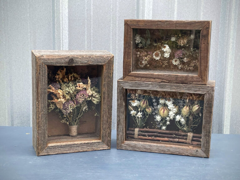 Three rustic Dried Floral Boxes display dried flowers—one upright with a bouquet, two stacked with floral arrangements behind glass—all set against a light, vertically striped wall.