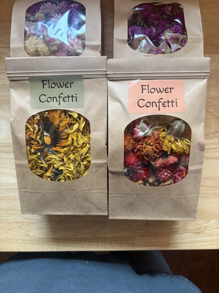 Four brown paper bags of Flower Confetti, each with a clear window showing vibrant dried flowers in yellow, orange, pink, and purple hues, arranged on a wooden surface.