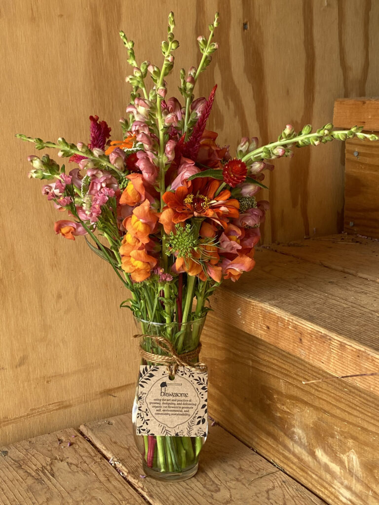 The Good Karma Bouquet, a vibrant mix of orange, red, and pink blooms in a glass jar with a twine-tied tag, is displayed on wooden steps against a wood-paneled background.